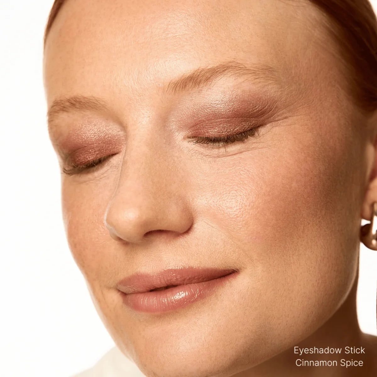 Close-up of a woman's face with closed eyes, wearing a beige eyeshadow stick labeled 'Cinnamon Spice'.