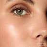 Close-up of a person's eye with a highlighter balm labeled 'Nude Glow' applied.