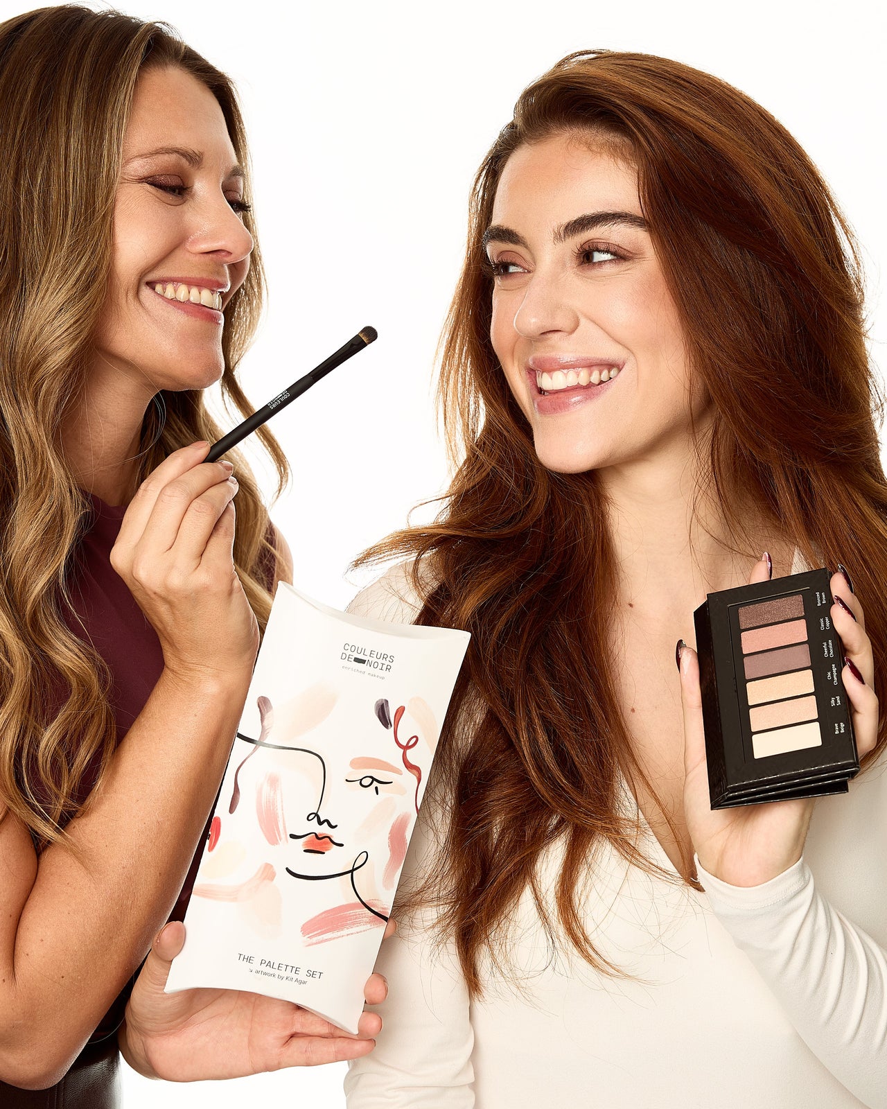 Two women holding makeup products and smiling on a white background